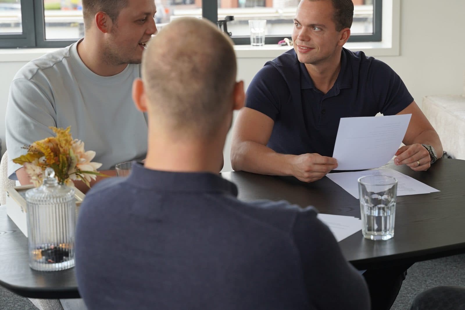 Twee collega's aan tafel op het kantoor van Elusive, één met een geprint vel in de hand, in gesprek met een derde die met de rug naar de camera zit