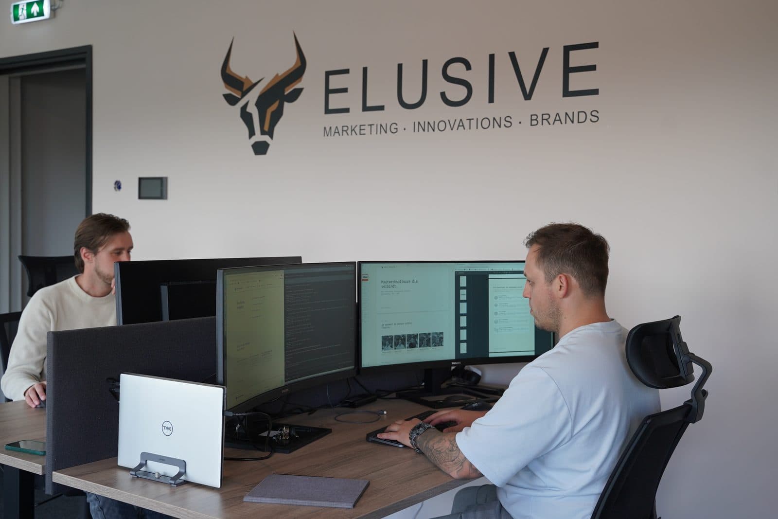 Twee mensen aan het werk aan bureaus met meerdere schermen, onder het logo van Elusive op de muur