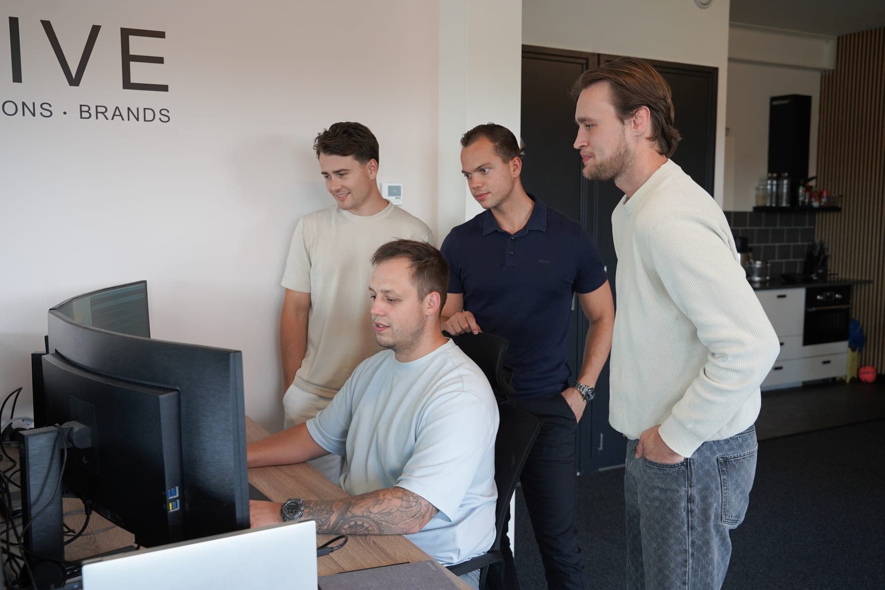 Four people at a desk in the Elusive office, three standing and looking over the shoulder of a colleague at his screen